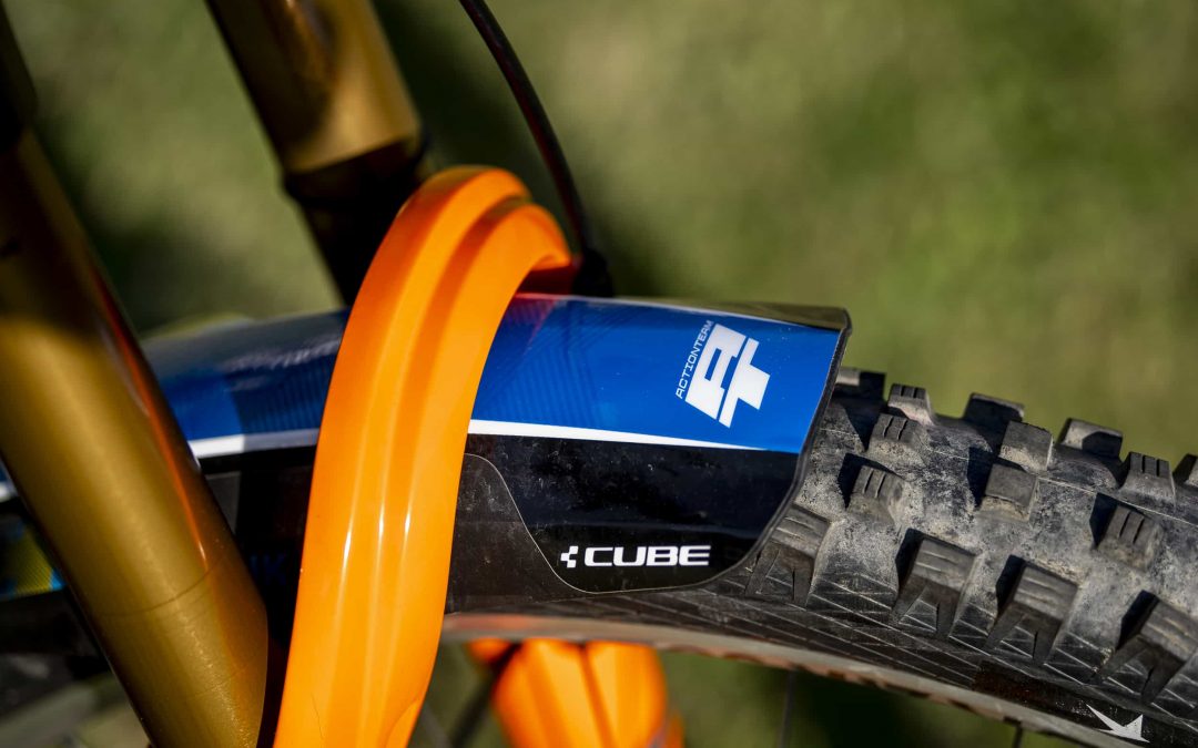 Nahaufnahme des Vorderreifens eines Mountainbikes mit einem CUBE-Logo auf dem Schutzblech und einer orangefarbenen Federgabel vor einem unscharfen grasbewachsenen Hintergrund.