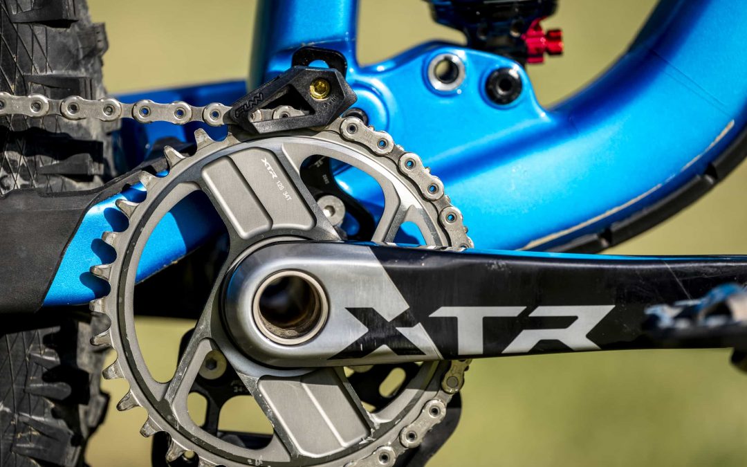 Nahaufnahme einer Mountainbike-Kurbelgarnitur und eines Kettenblatts mit einem Shimano XTR-Kurbelarm, die an einem blauen Fahrradrahmen befestigt sind. Das Bild zeigt detaillierte Fahrradkomponenten und einen Teil des Hinterreifens.