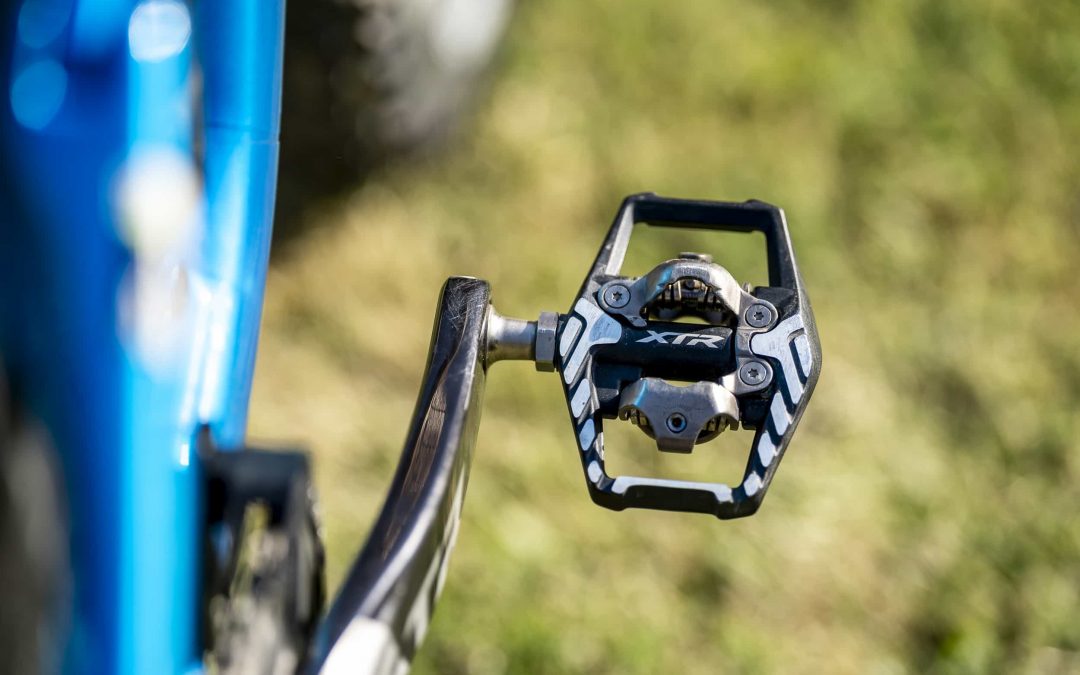 Nahaufnahme eines schwarzen Shimano XTR-Mountainbike-Pedals, das an einem Kurbelarm befestigt ist, wobei ein Teil eines blauen Fahrradrahmens vor einem unscharfen grasbewachsenen Hintergrund zu sehen ist.