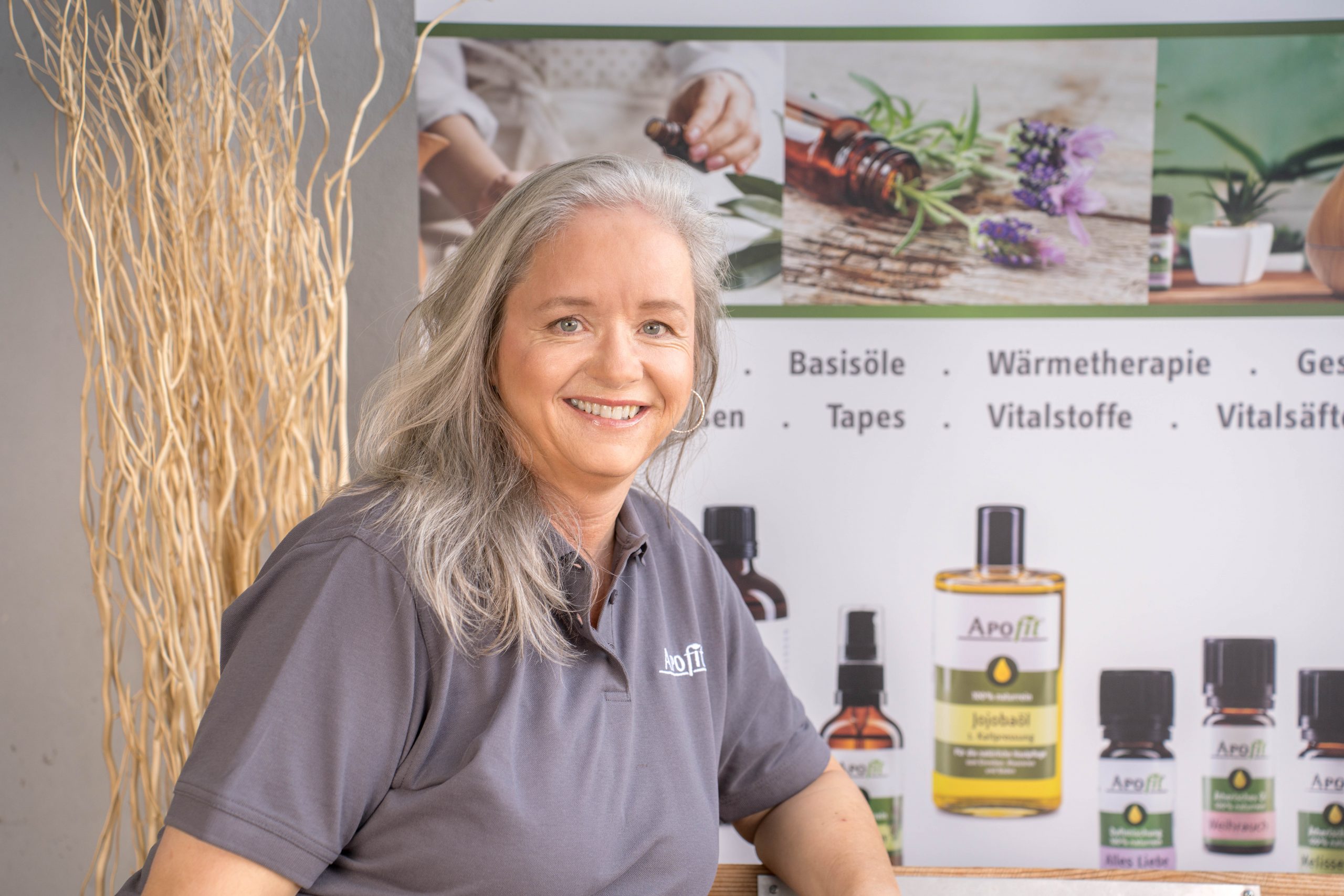 Eine lächelnde Frau mit langen grauen Haaren und einem grauen Polohemd sitzt vor einer Auslage mit ätherischen Ölen und Wellnessprodukten. Neben ihr sind getrocknete dekorative Zweige zu sehen.