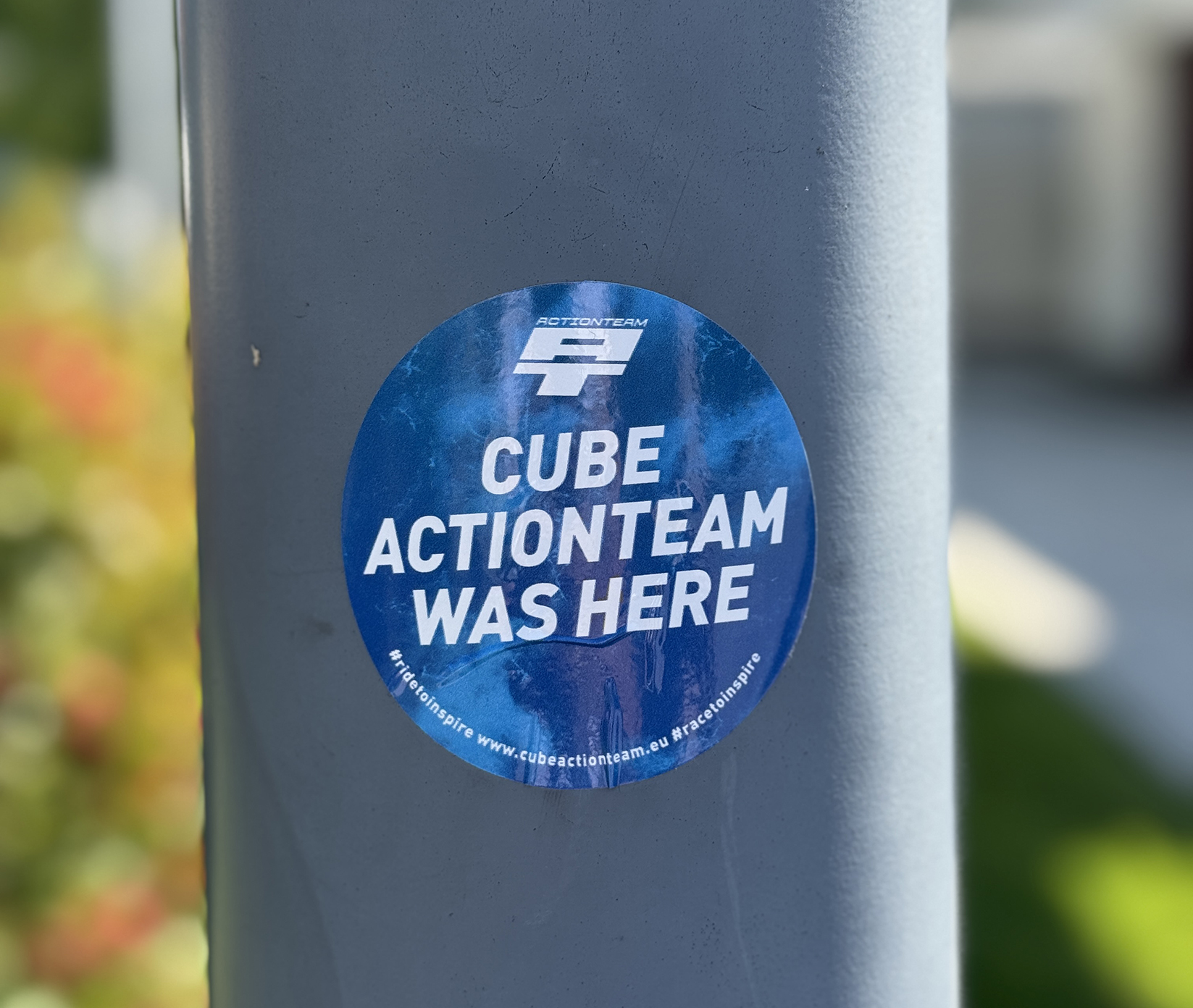 Ein blauer kreisförmiger Aufkleber an einem Pfahl zeigt die Aufschrift CUBE ACTIONTEAM WAS HERE mit einem Logo und einem Link zur Website am unteren Rand. Der Hintergrund besteht aus verschwommenem Grün.