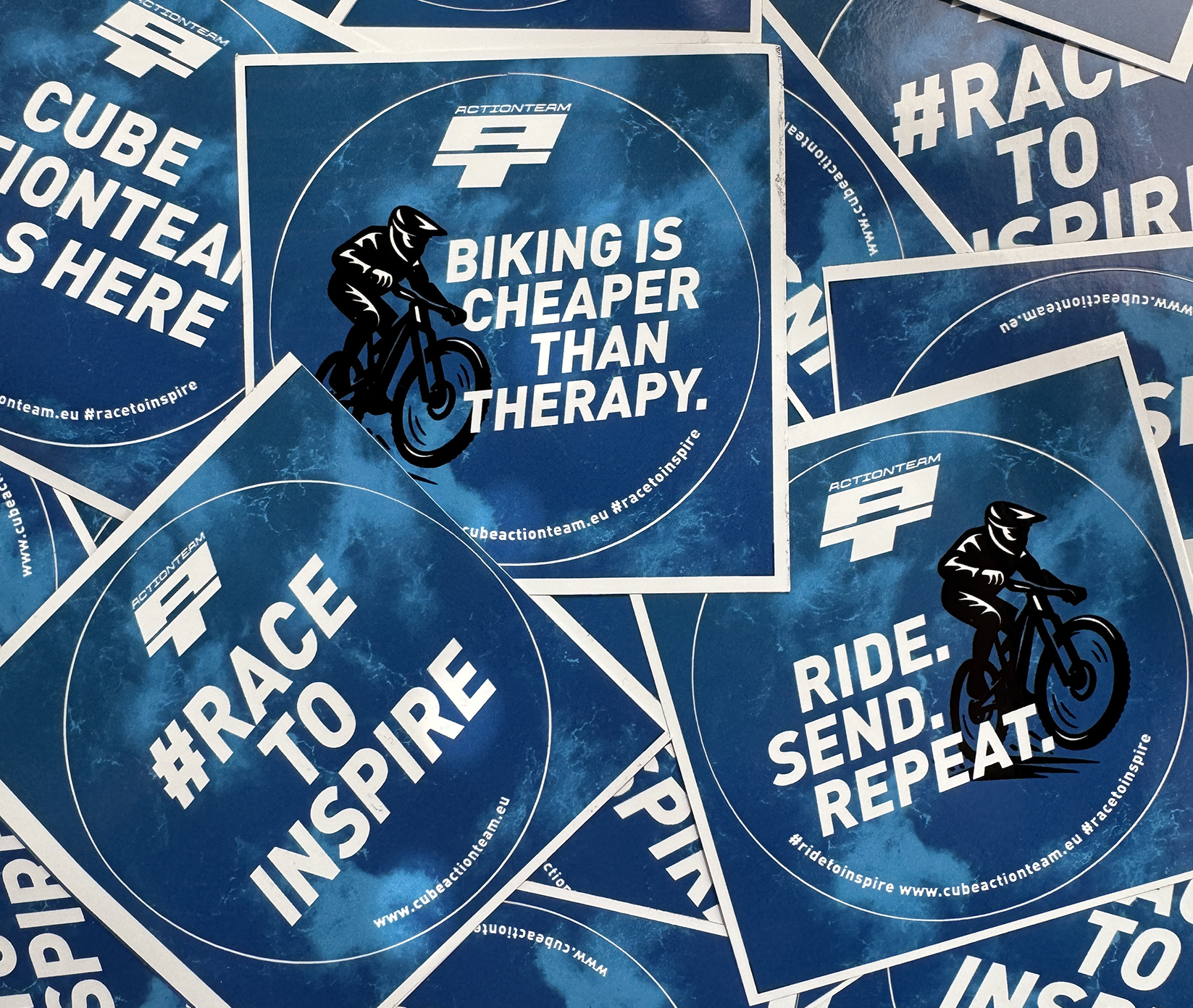 Blaue Aufkleber mit motivierenden Radfahrersprüchen wie "BIKING IS CHEAPER THANAPY", "RACE TO INSPIRE" und "RIDE. SENDEN. REPEAT.", zusammen mit Abbildungen von Radfahrern und Logos des Cube Action Teams.