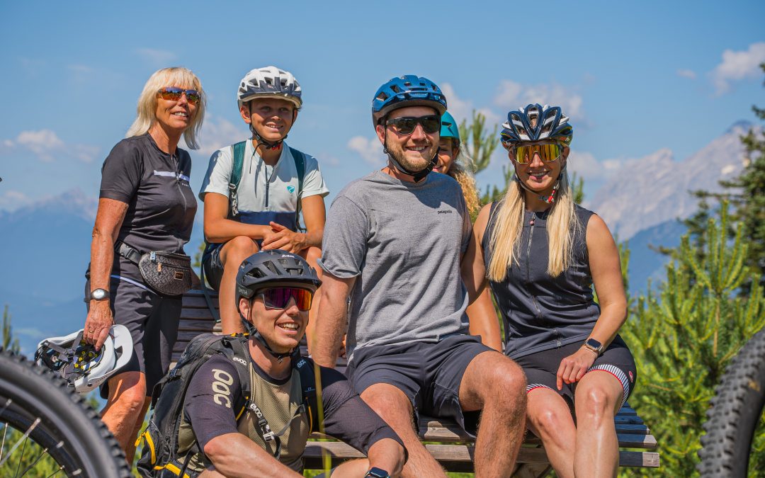 Eine Gruppe von sechs Personen, die Fahrradkleidung und Helme tragen, lächeln, während sie an einem sonnigen Tag im Freien posieren, umgeben von Grün und Bergen im Hintergrund.