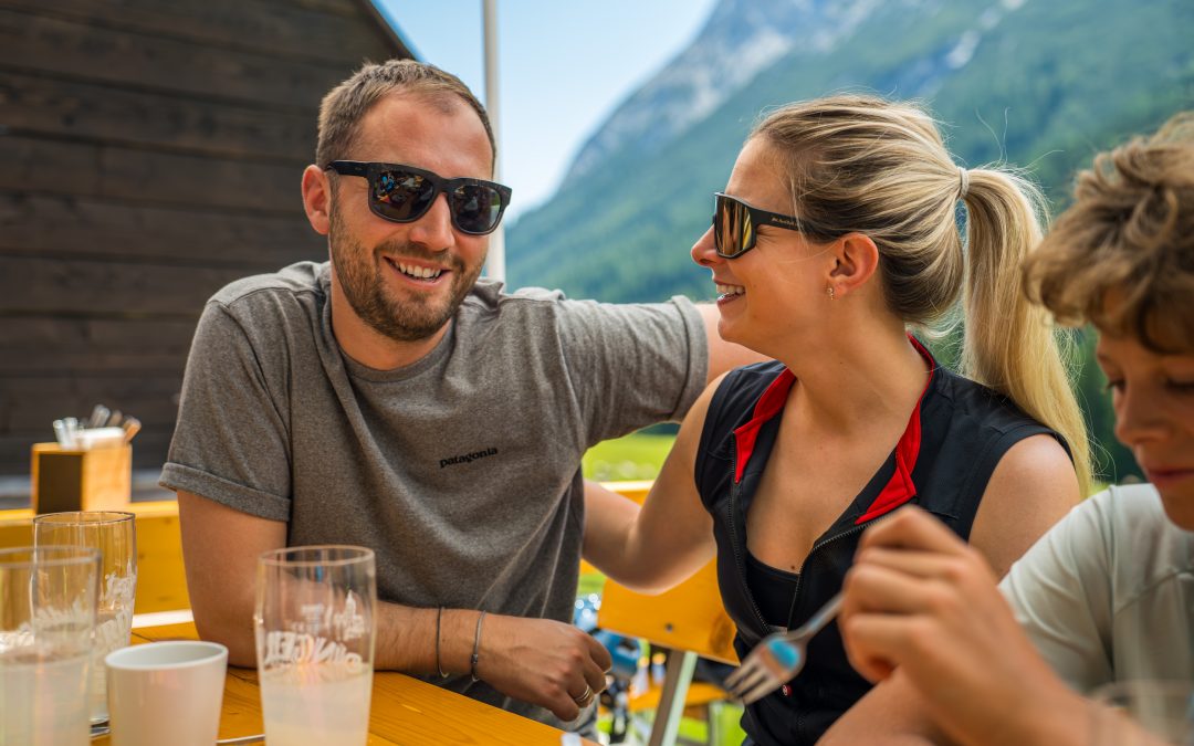 Ein Mann und eine Frau mit Sonnenbrille lächeln und sitzen dicht beieinander an einem Tisch im Freien, mit Bergen und Grünflächen im Hintergrund. Eine weitere Person ist teilweise im Vordergrund zu sehen.