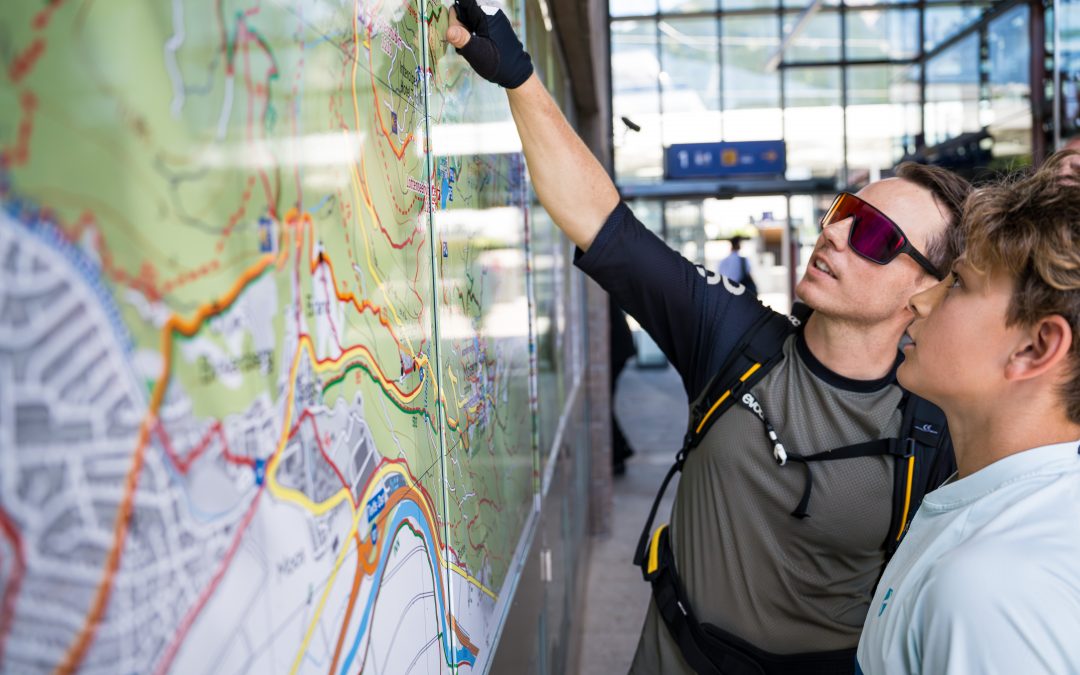 Zwei Personen studieren eine große Karte, die an einer Station ausgestellt ist, wobei eine Person auf einen Ort zeigt. Beide tragen legere Outdoor-Kleidung, und das Sonnenlicht fällt durch Glasscheiben über ihnen.