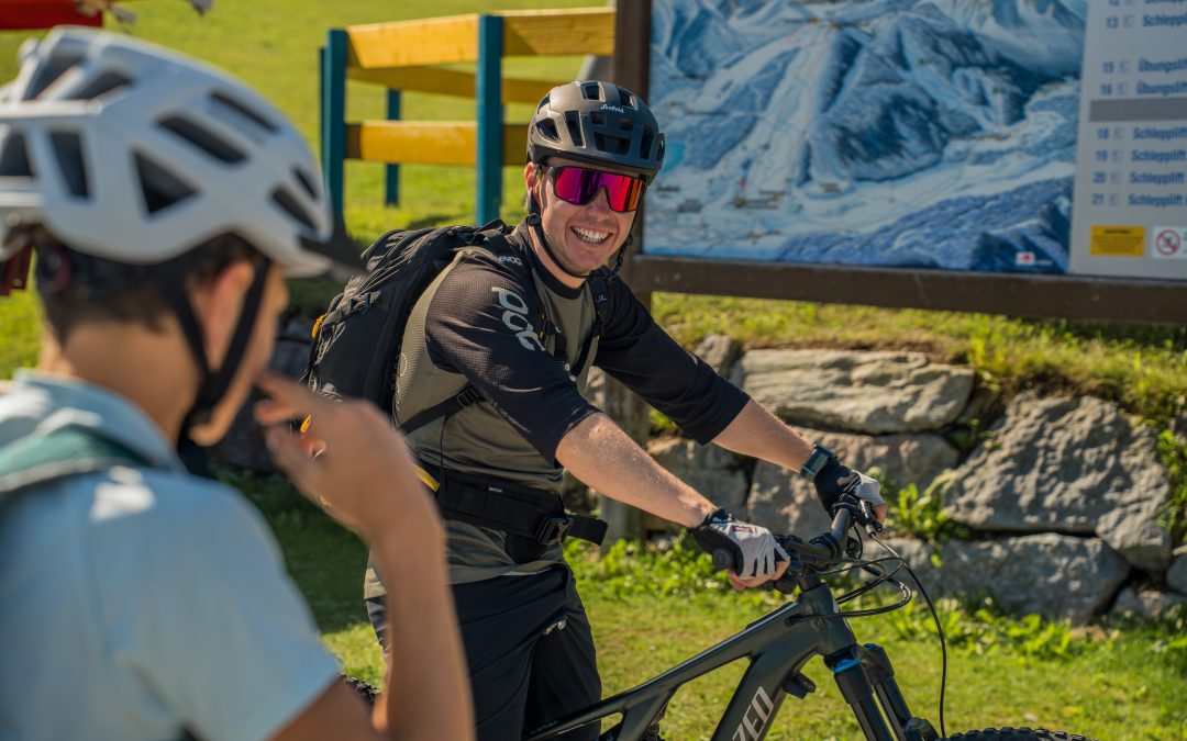 Ein lächelnder Radfahrer mit Helm, Sonnenbrille und Rucksack sitzt auf einem Mountainbike im Freien, einem anderen Radfahrer gegenüber, mit einer Wanderkarte und einem grasbewachsenen Hügel im Hintergrund.