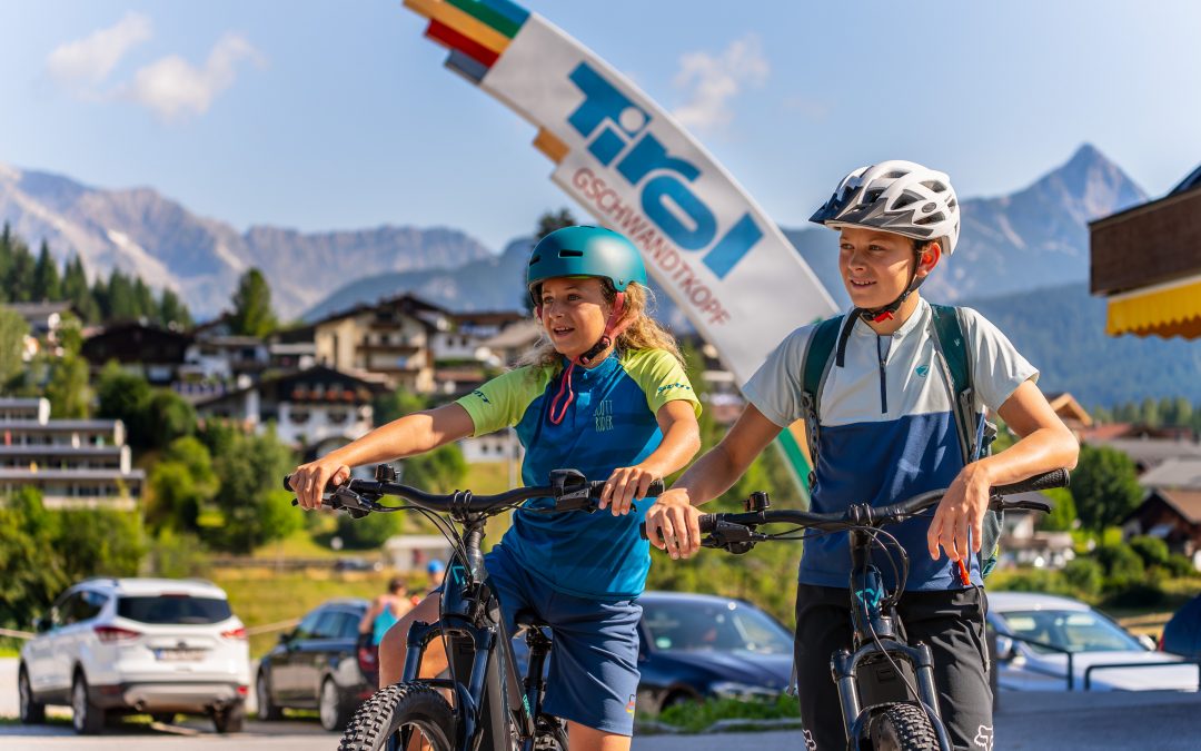 Zwei lächelnde, helmtragende Kinder fahren in einem sonnigen Bergdorf mit dem Fahrrad. Im Hintergrund sind Häuser, Autos und ein buntes Tirol-Schild zu sehen, dahinter Berge und blauer Himmel.