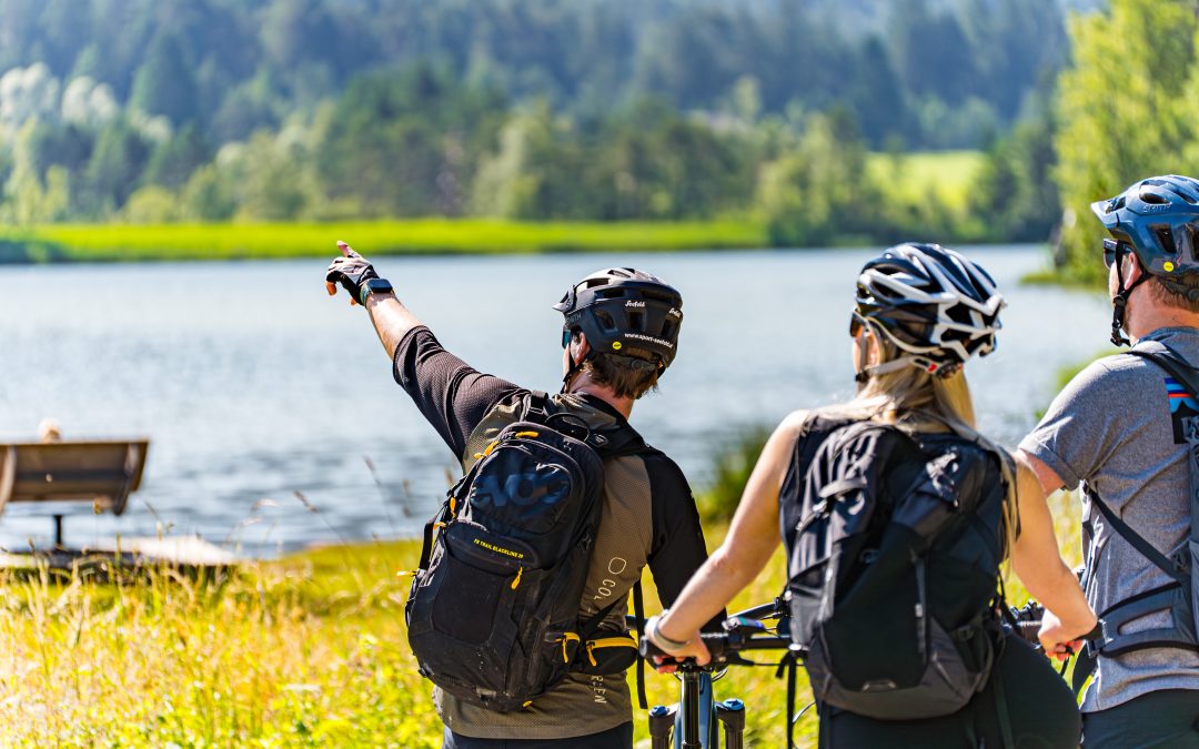 Drei Radfahrer mit Helmen und Rucksäcken stehen mit ihren Fahrrädern an einem See, wobei eine Person auf etwas in der Ferne auf der anderen Seite des Wassers deutet, umgeben von üppigem Grün und Bäumen.