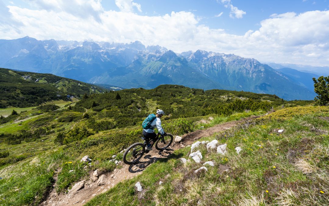 Ein Mountainbiker fährt einen gewundenen Feldweg hinunter, umgeben von üppig grünen Hügeln, mit hohen, schroffen Bergen und einem teilweise bewölkten Himmel im Hintergrund.