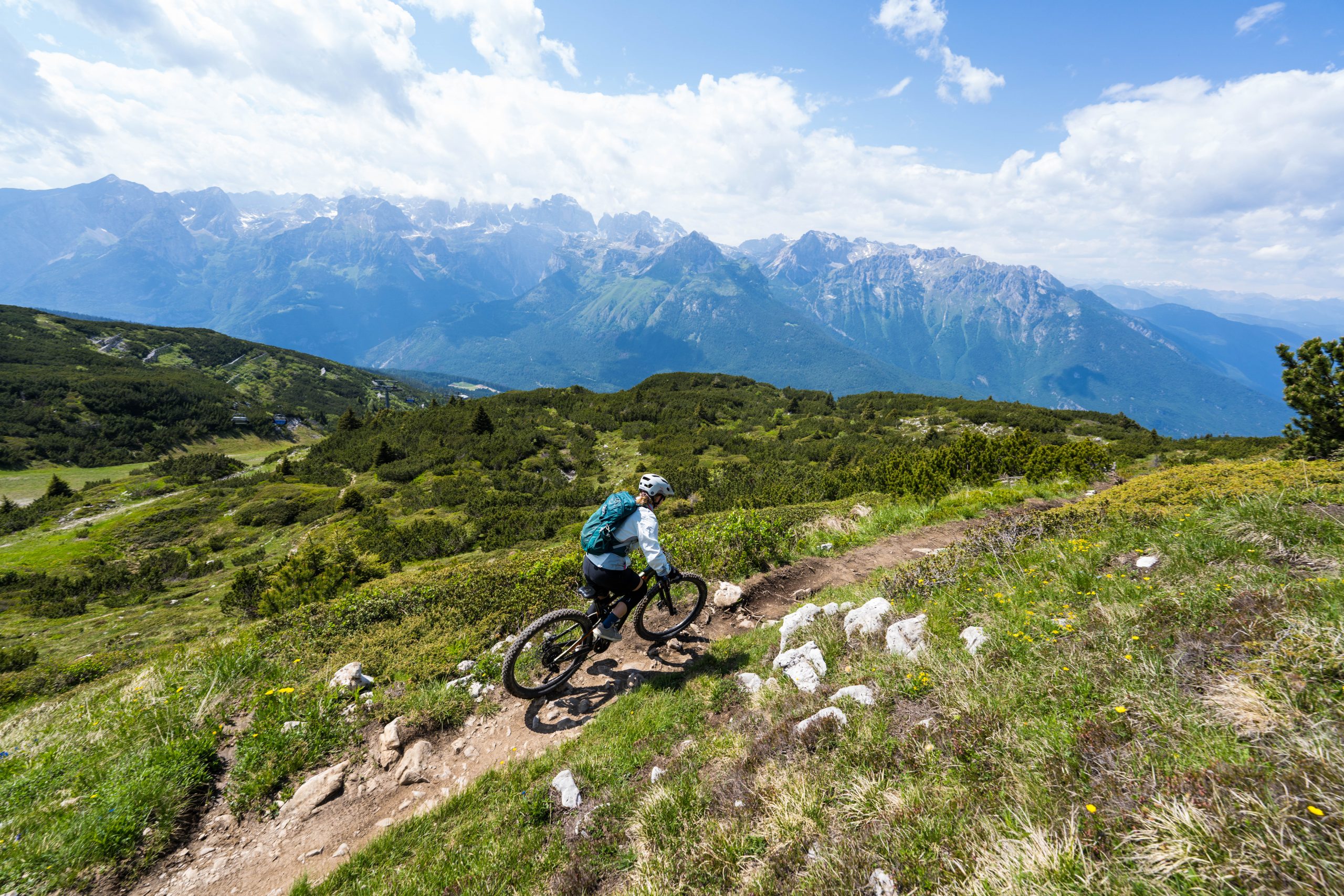 Ein Mountainbiker fährt einen gewundenen Feldweg hinunter, umgeben von üppig grünen Hügeln, mit hohen, schroffen Bergen und einem teilweise bewölkten Himmel im Hintergrund.