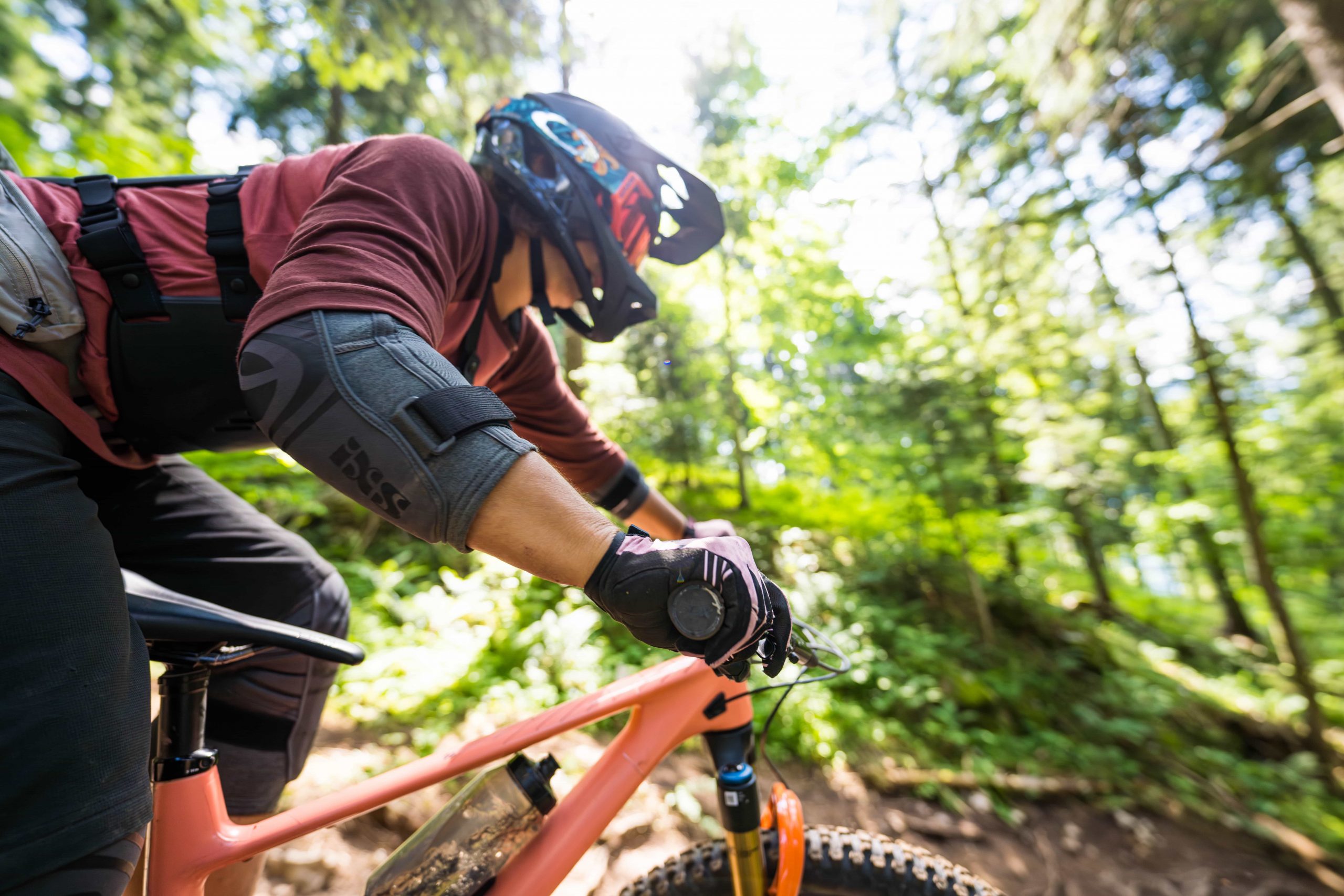 Eine Person mit Schutzausrüstung und Helm fährt auf einem Mountainbike durch einen sonnenbeschienenen Waldweg, hält sich am Lenker fest und schaut nach vorne.