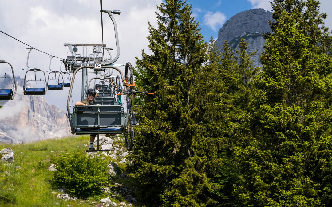 Eine Person fährt mit einem Skilift und einem Mountainbike auf einen grünen, bewaldeten Berg mit hohen Kiefern und felsigen Gipfeln im Hintergrund unter einem teilweise bewölkten Himmel.