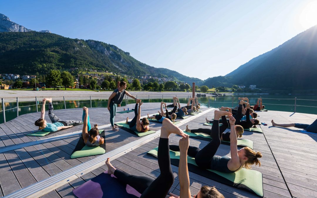 Eine Gruppe von Menschen praktiziert Yoga auf Matten auf einer Holzterrasse an einem See, umgeben von Bergen und Grün, unter strahlend blauem Himmel, angeleitet von einem Lehrer.