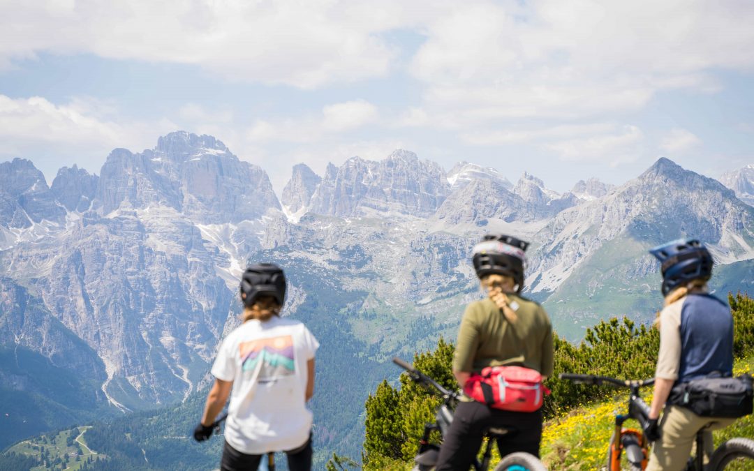 Drei Radfahrer mit Helmen und Outdoor-Ausrüstung machen mit ihren Fahrrädern eine Pause und blicken auf eine malerische Berglandschaft mit schroffen, schneebedeckten Gipfeln unter einem teilweise bewölkten Himmel.