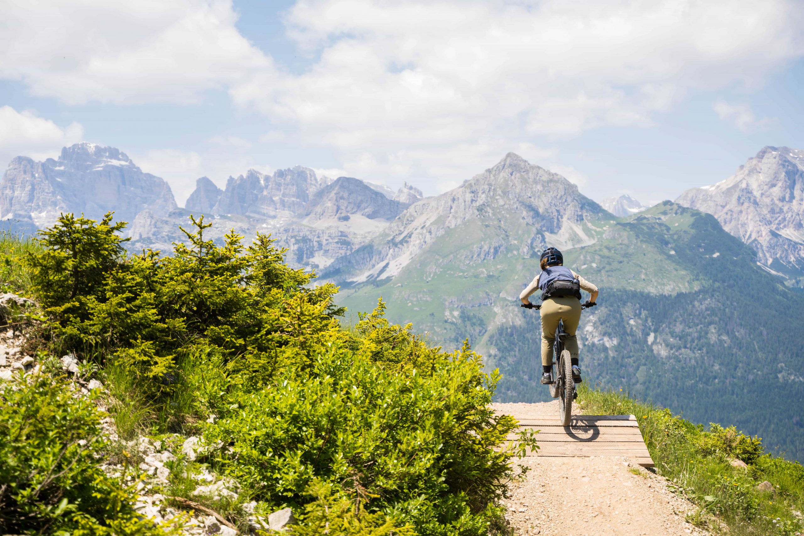 Eine Person fährt mit dem Mountainbike auf einem von grünen Büschen umgebenen Weg mit schroffen, schneebedeckten Bergen und einem teilweise bewölkten Himmel im Hintergrund.