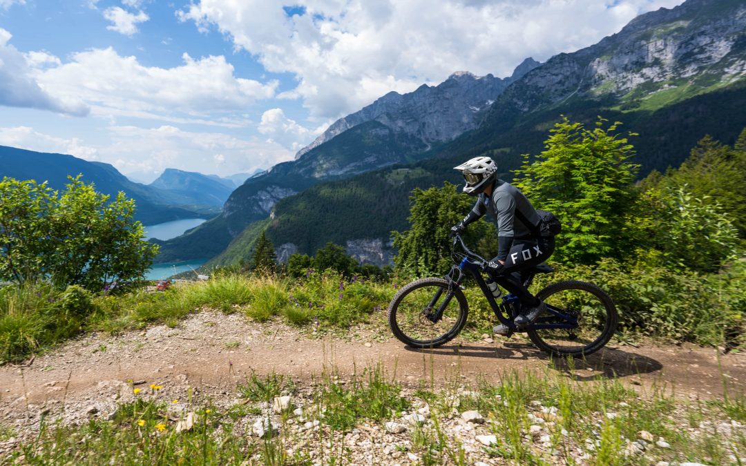 Ein Radfahrer mit Helm und Ausrüstung fährt mit einem Mountainbike auf einem unbefestigten Weg, umgeben von grünen Bäumen und Gras, mit Bergen und einem blauen See im Hintergrund unter einem teilweise bewölkten Himmel.
