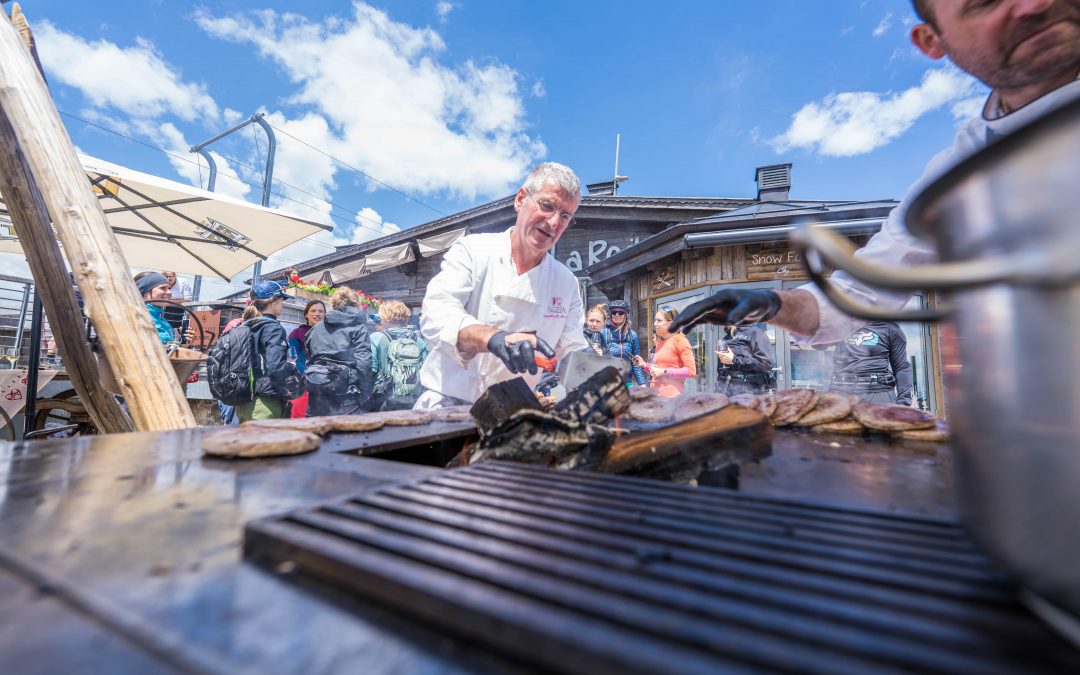 Ein Koch grillt auf einem belebten Markt mit einer Zange Lebensmittel im Freien, wobei Rauch vom Grill aufsteigt. Menschen mit Rucksäcken versammeln sich um ihn herum, und über ihm ist ein blauer Himmel mit Wolken zu sehen.