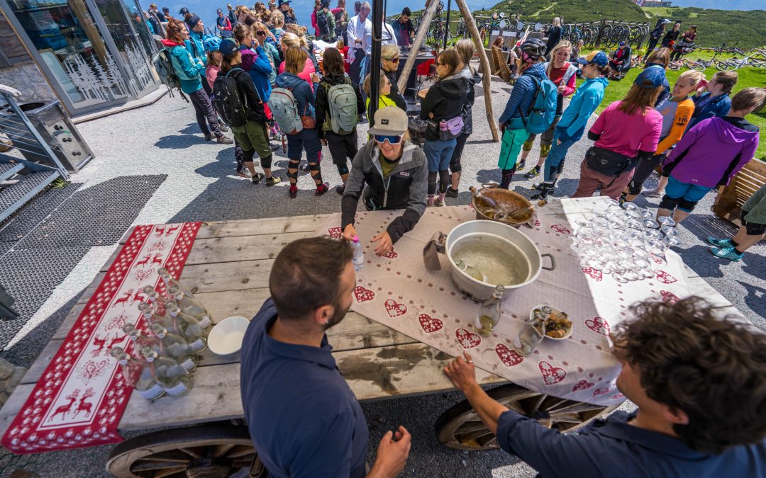 Eine Gruppe von Menschen versammelt sich im Freien an einem Holztisch mit Getränken und Snacks, während sich zwei Kellner mit einer Frau unterhalten. Im Hintergrund sind die Berge zu sehen, und die meisten Menschen tragen Wanderkleidung und Rucksäcke.