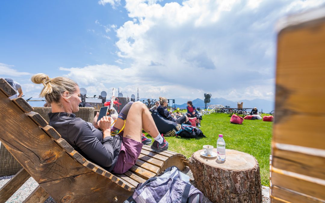 Eine Frau entspannt sich auf einem hölzernen Liegestuhl im Freien, in der Hand einen Snack. Andere Menschen und Rucksäcke liegen auf dem Gras in der Nähe. Eine Tasse, ein Teller und eine Wasserflasche stehen auf einem Baumstumpf. Im Hintergrund sind Berge und ein bewölkter Himmel zu sehen.