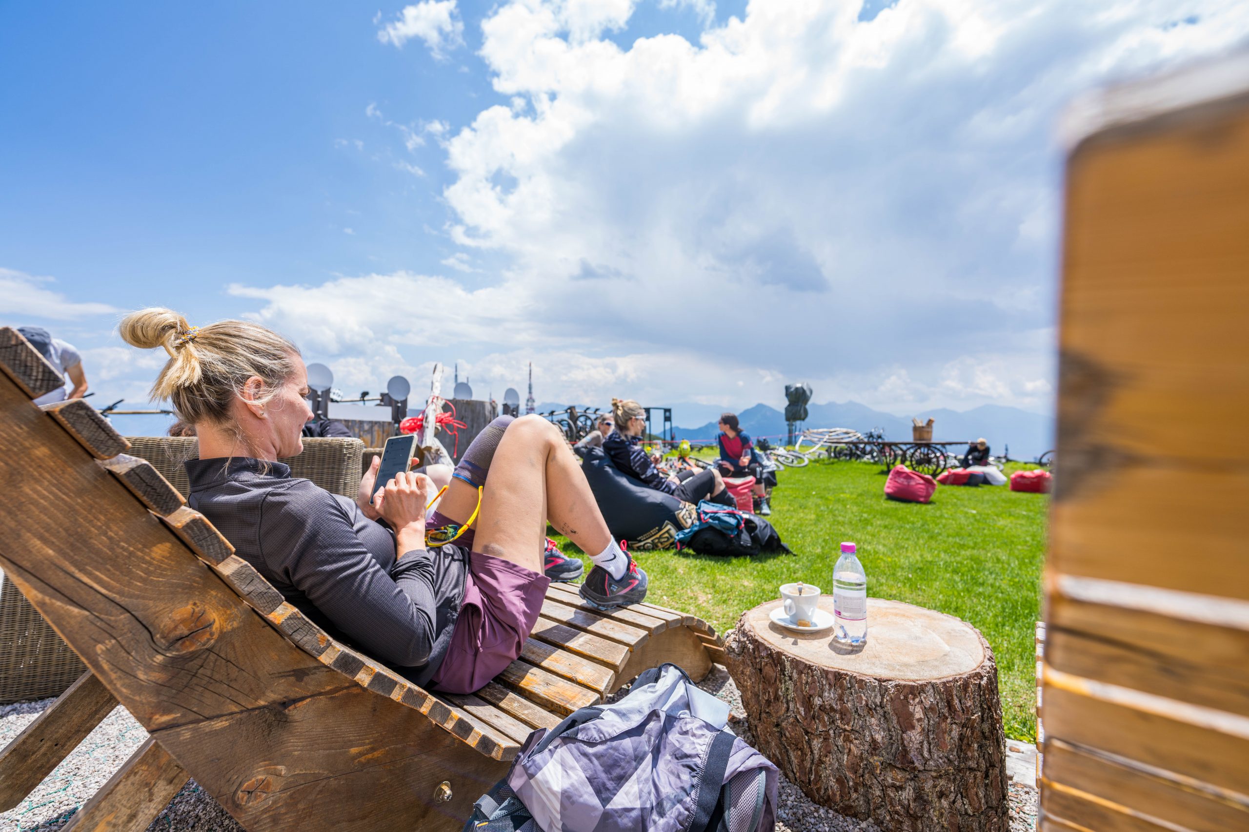 Eine Frau entspannt sich auf einem hölzernen Liegestuhl im Freien, in der Hand einen Snack. Andere Menschen und Rucksäcke liegen auf dem Gras in der Nähe. Eine Tasse, ein Teller und eine Wasserflasche stehen auf einem Baumstumpf. Im Hintergrund sind Berge und ein bewölkter Himmel zu sehen.