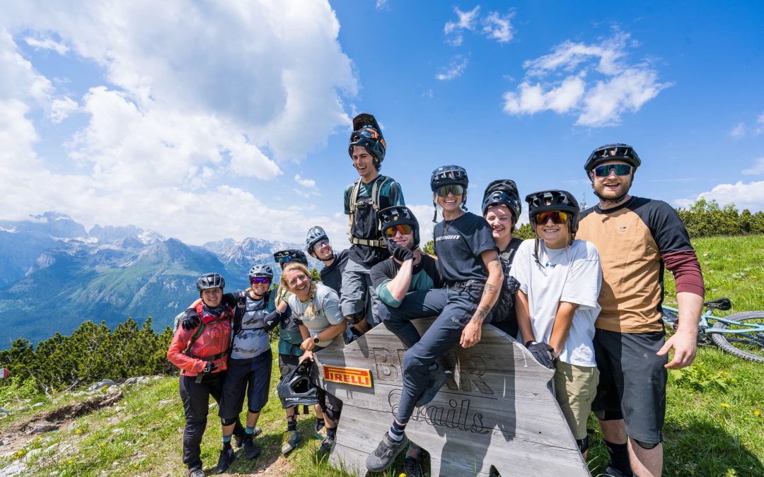 Eine Gruppe von Radfahrern mit Helmen und Ausrüstung posiert zusammen auf einem grasbewachsenen Berghang mit Fahrrädern in der Nähe, lächelnd vor einem Holzschild, mit malerischen Bergen und blauem Himmel im Hintergrund.