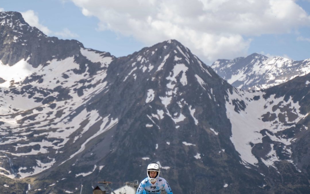 Eine Person, die einen Helm trägt, fährt mit dem Fahrrad auf einem Schotterweg in einer bergigen Gegend mit Schneeflecken auf den Gipfeln und einem teilweise bewölkten Himmel.