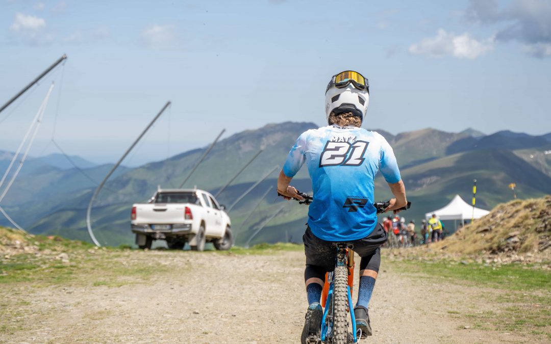 Ein Radfahrer mit blauem Trikot und Helm fährt mit seinem Mountainbike auf einem unbefestigten Weg in Richtung einer bergigen Landschaft, in der ein weißer Lastwagen und Zelte in der Ferne unter einem teilweise bewölkten Himmel zu sehen sind.