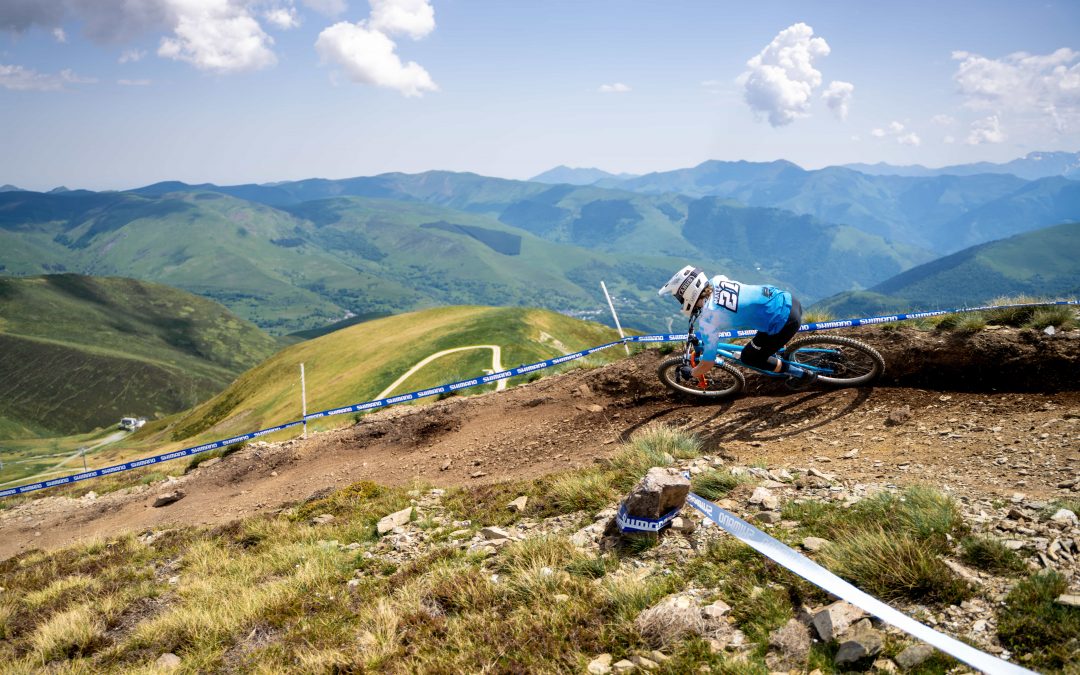 Ein Mountainbiker im blauen Outfit fährt auf einem unbefestigten Weg an einem Hang, umgeben von grünen Bergen und einem blauen Himmel mit vereinzelten Wolken. Weiße Absperrungen säumen den kurvenreichen Weg.
