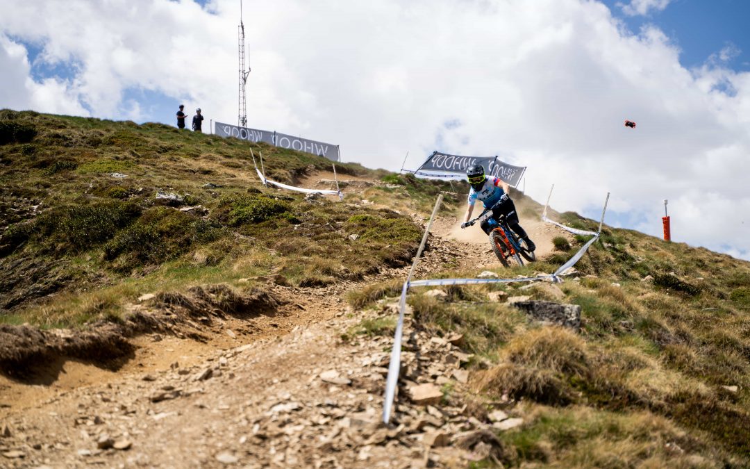 Ein Mountainbiker fährt bei teilweise bewölktem Himmel einen felsigen Pfad an einem Hang hinunter. Drei Personen stehen auf dem Gipfel in der Nähe von Absperrungen und einem Funkturm. Grasflächen säumen den zerklüfteten Feldweg.