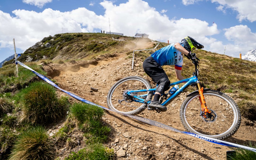Ein Mountainbiker in einem blauen Trikot und mit Helm fährt einen steinigen, unbefestigten Weg an einem Berghang hinunter, der Staub aufwirbelt, wobei Gras und Klebeband die Strecke unter einem teilweise bewölkten Himmel markieren.