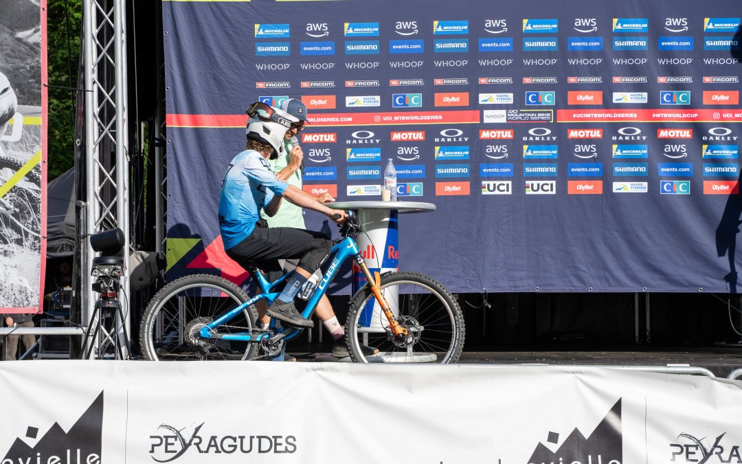 Ein Radfahrer mit Helm und blauer Kleidung sitzt auf einem Mountainbike auf einer Bühne vor einer Kulisse mit Logos bei einer Weltcup-Veranstaltung in Loudenvielle, Frankreich.