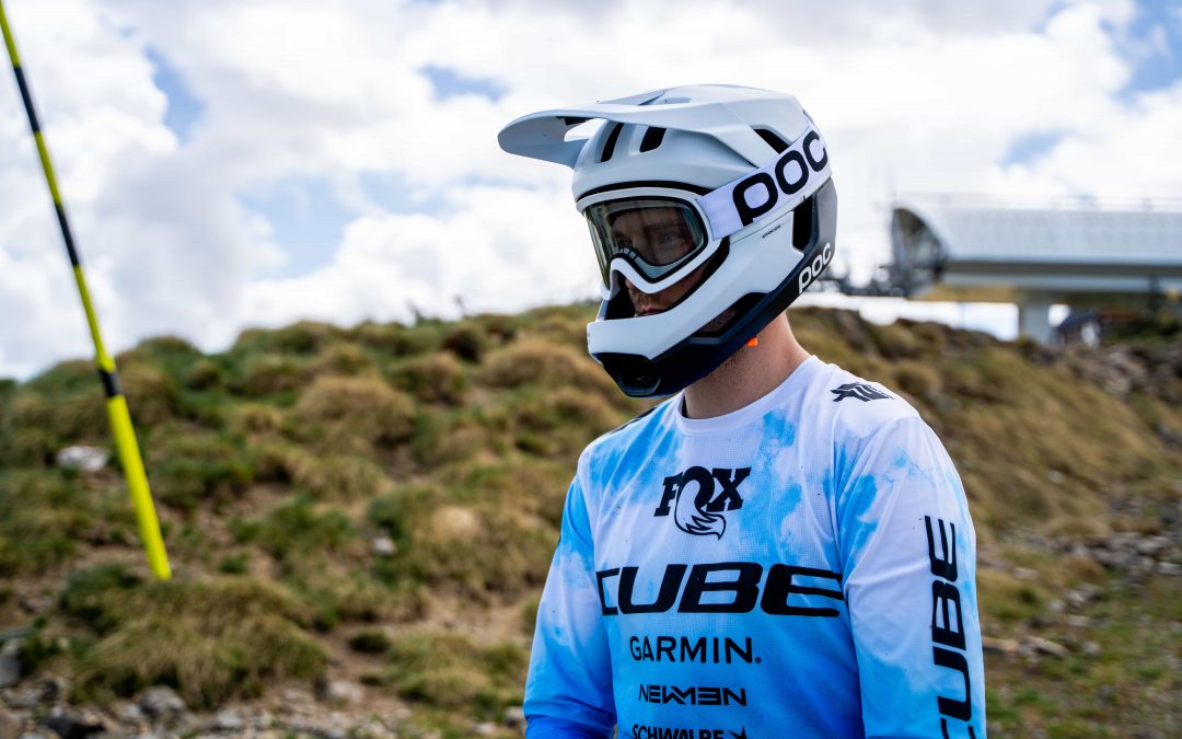 Eine Person, die einen weißen Integralhelm, eine Schutzbrille und ein blau-weißes Motocross-Trikot trägt, steht im Freien auf einem grasbewachsenen, felsigen Hügel mit einem bewölkten Himmel im Hintergrund.