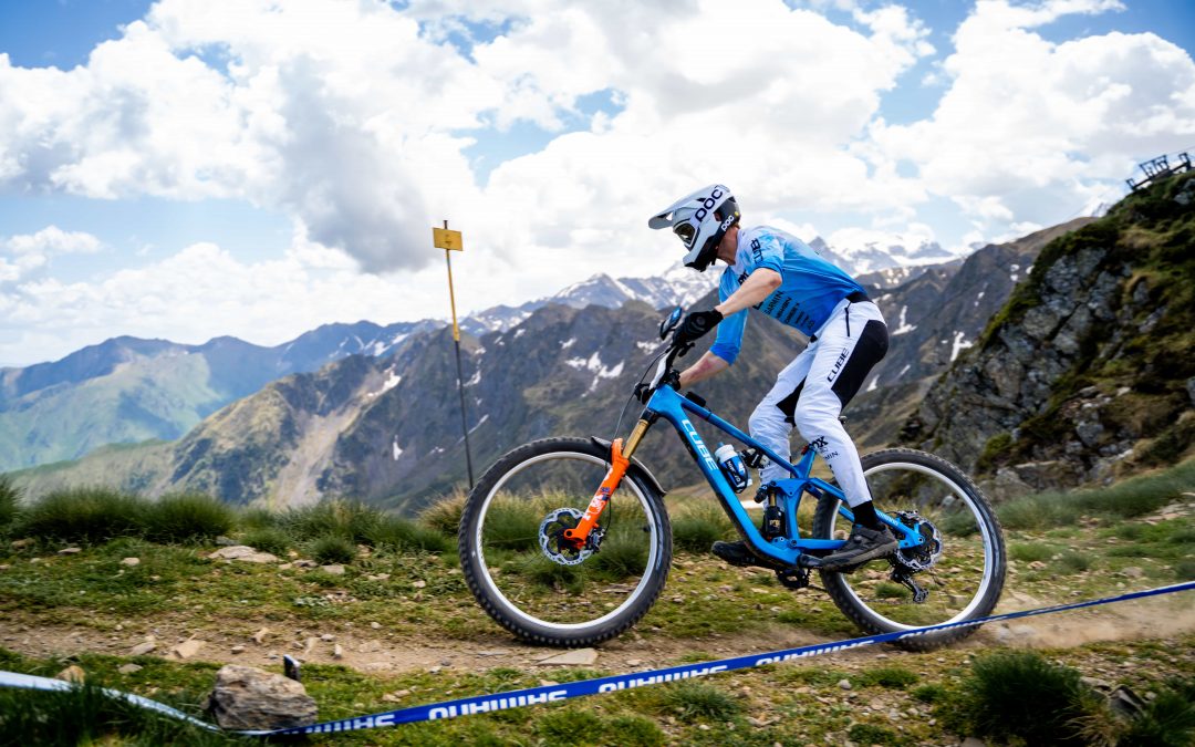 Ein Mountainbiker in blau-weißer Kleidung fährt bergauf auf einem felsigen Weg mit Bergen und einem teilweise bewölkten Himmel im Hintergrund. Gras und Felsen säumen den Weg, und in der Nähe ist eine Streckenmarkierung zu sehen.