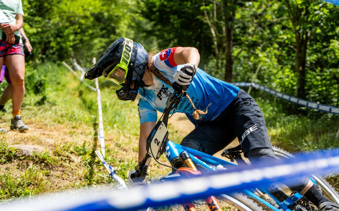Ein Mountainbiker in voller Montur und mit Helm biegt während eines Rennens auf einem Waldweg scharf in eine Kurve ein, wobei die Strecke mit blauem und weißem Band markiert ist und im Hintergrund Bäume zu sehen sind.
