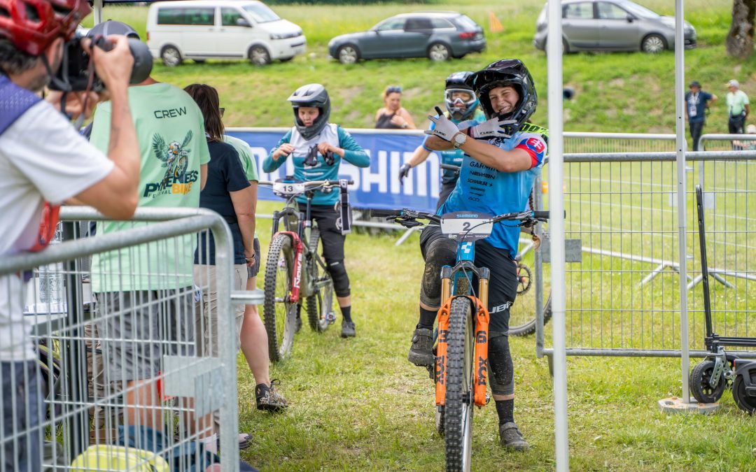 Ein Mountainbiker in blauer Kleidung und mit Helm lächelt und zeigt in die Kamera, während er sein Fahrrad im Zielbereich eines Rennens hält. Im Hintergrund sind Mitarbeiter der Veranstaltung und Zuschauer zu sehen.
