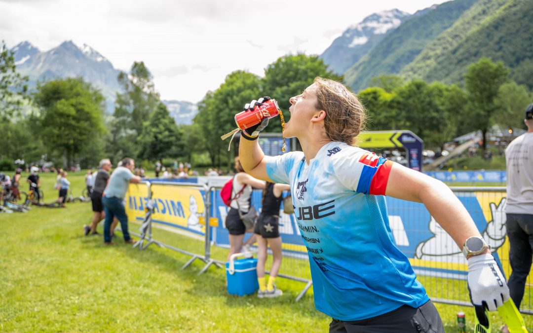 Ein Radfahrer in einem blau-weißen Trikot trinkt aus einer Flasche, während er neben einer Rennstrecke läuft, mit Bergen, Bäumen und Zuschauern im Hintergrund an einem sonnigen Tag.