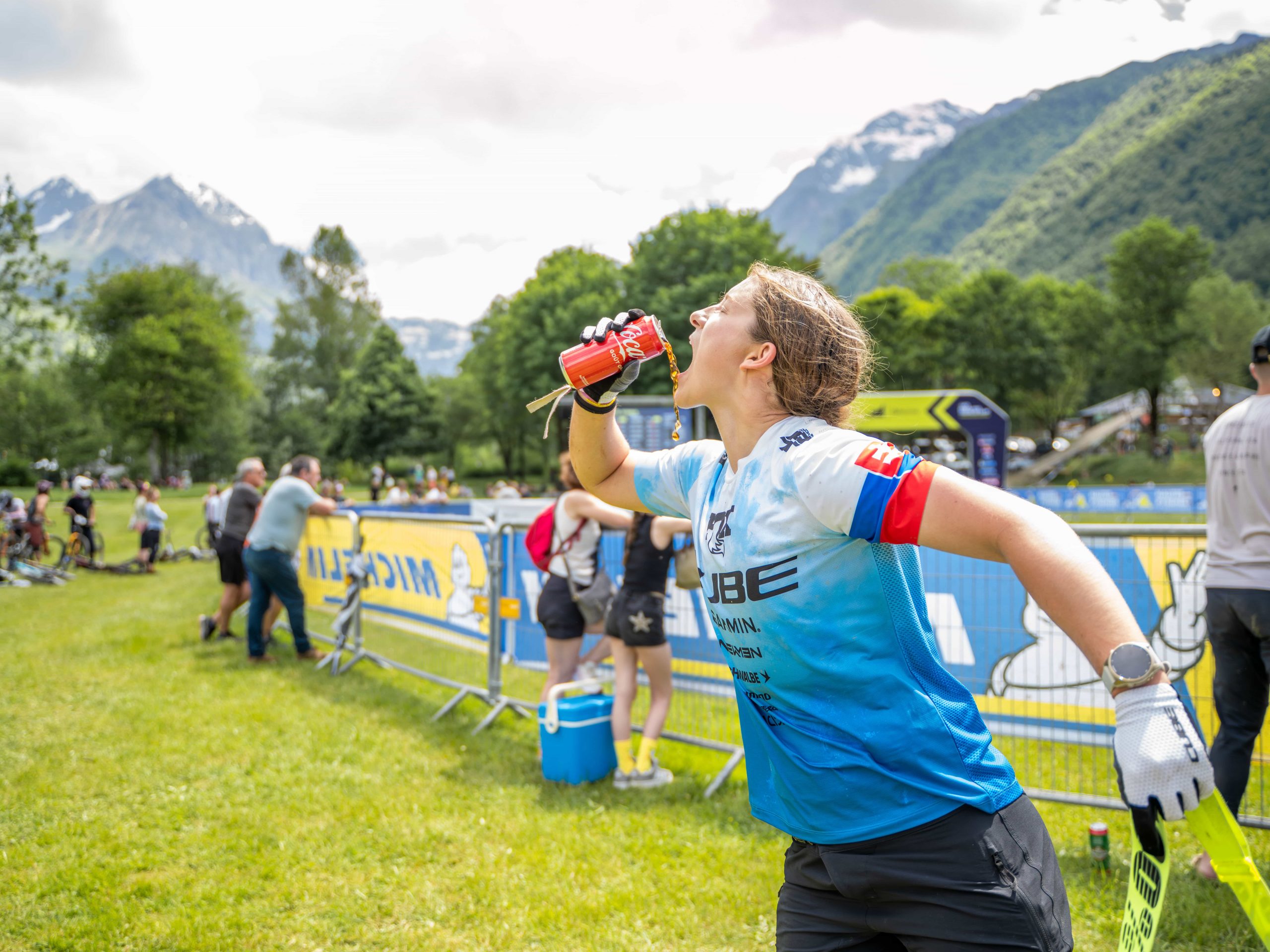 Ein Radfahrer in einem blau-weißen Trikot trinkt aus einer Flasche, während er neben einer Rennstrecke läuft, mit Bergen, Bäumen und Zuschauern im Hintergrund an einem sonnigen Tag.