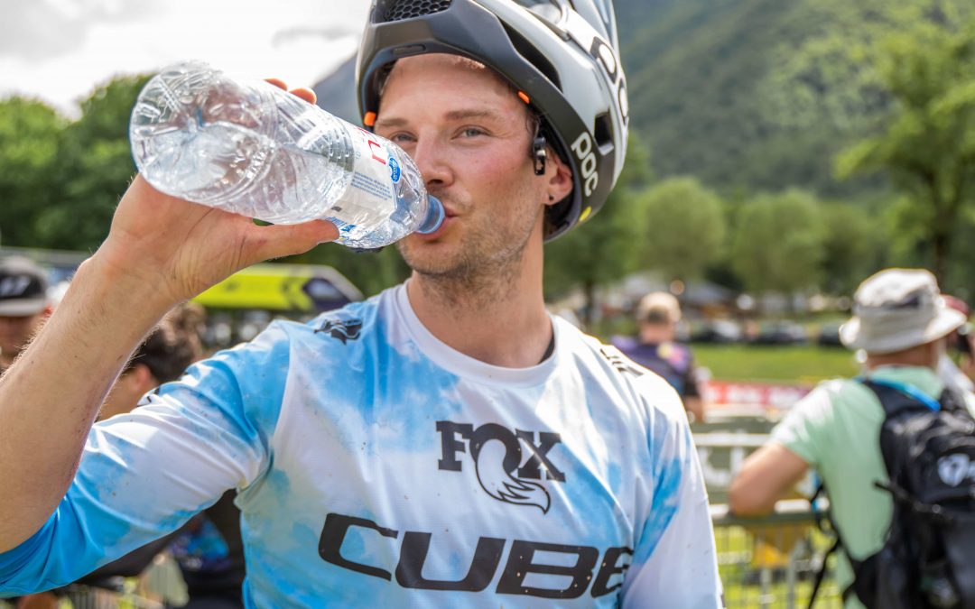 Ein Mountainbiker, der einen Helm und ein blaues FOX CUBE-Trikot trägt, trinkt Wasser aus einer Flasche und hält eine Dose Coca-Cola. Er befindet sich im Freien bei einer Veranstaltung, mit Menschen und grünen Hügeln im Hintergrund.