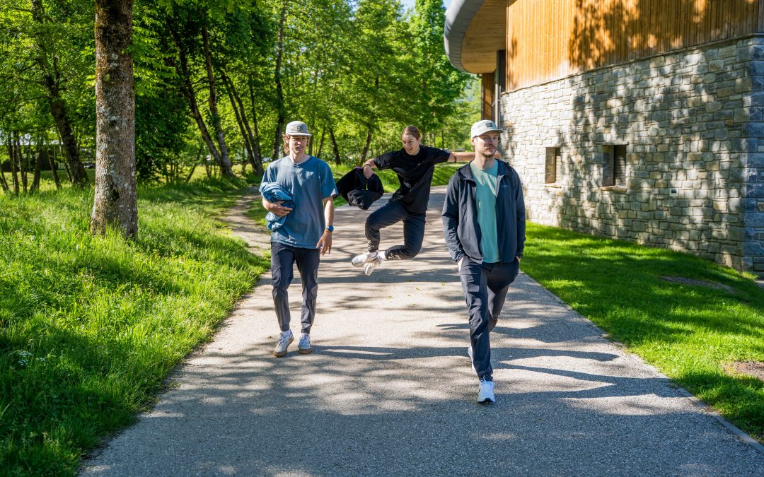Drei junge Männer gehen auf einem sonnigen Weg neben einem steinernen Gebäude, wobei ein Mann in der Mitte spielerisch springt, während die anderen ruhig gehen. Üppige grüne Bäume und Gras umgeben sie.