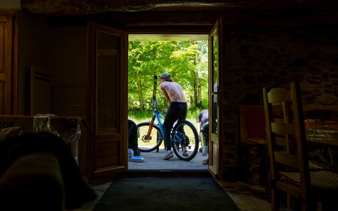 Eine Person ohne Hemd steht vor einem Haus und stützt sich auf ein blaues Fahrrad, das von innen durch eine offene Holztür betrachtet wird. Im Hintergrund sind Sonnenlicht und Grünflächen zu sehen.