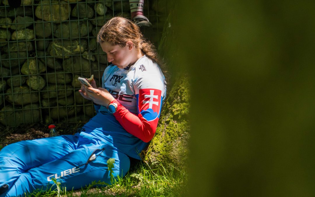 Eine Person in blau-weißer Sportkleidung mit einem Aufnäher mit slowakischer Flagge sitzt im Gras, lehnt sich an eine mit Steinen gefüllte Metallkonstruktion und schaut auf ihr Telefon. Eine weitere Person ist teilweise hinter der Struktur zu sehen.