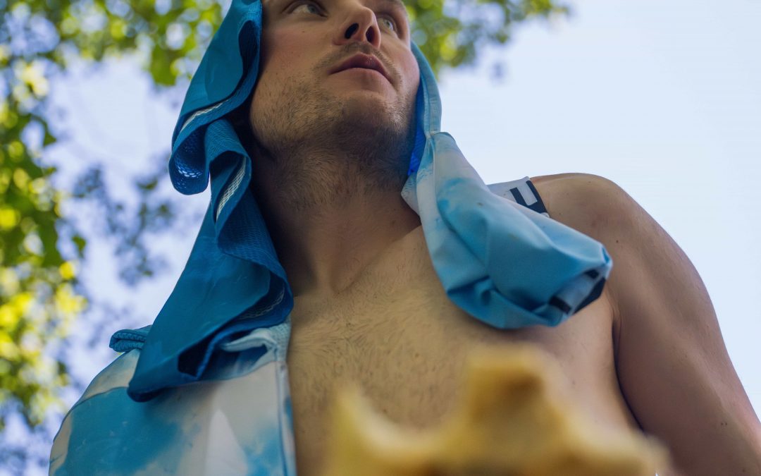 Eine Person ohne Hemd mit einem blau-weißen Handtuch über dem Kopf blickt in die Ferne, während sie ein teilweise gegessenes Sandwich in der Hand hält. Grüne Blätter und blauer Himmel sind darüber zu sehen.
