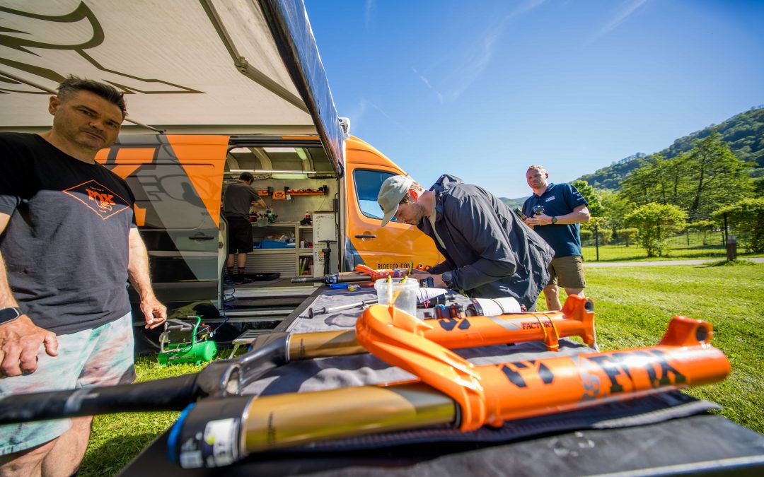 Drei Männer arbeiten an einer mobilen Fahrradreparaturstation, die neben einem orangefarbenen Lieferwagen auf einer Wiese im Freien unter blauem Himmel aufgebaut ist. Auf einem Tisch im Vordergrund liegen Federgabeln und Werkzeuge für Fahrräder.
