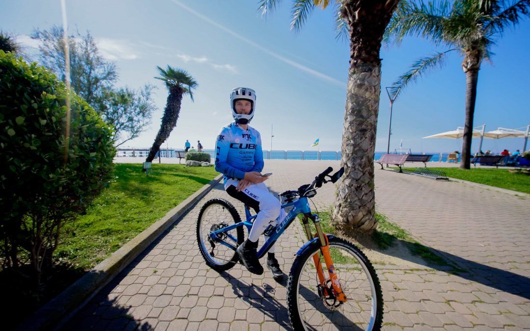 Ein Radfahrer in blau-weißer Kleidung sitzt auf einem Mountainbike auf einem sonnigen, palmengesäumten Weg in der Nähe des Strandes, mit grünen Büschen, Bänken und Menschen, die sich unter Sonnenschirmen entspannen, im Hintergrund.