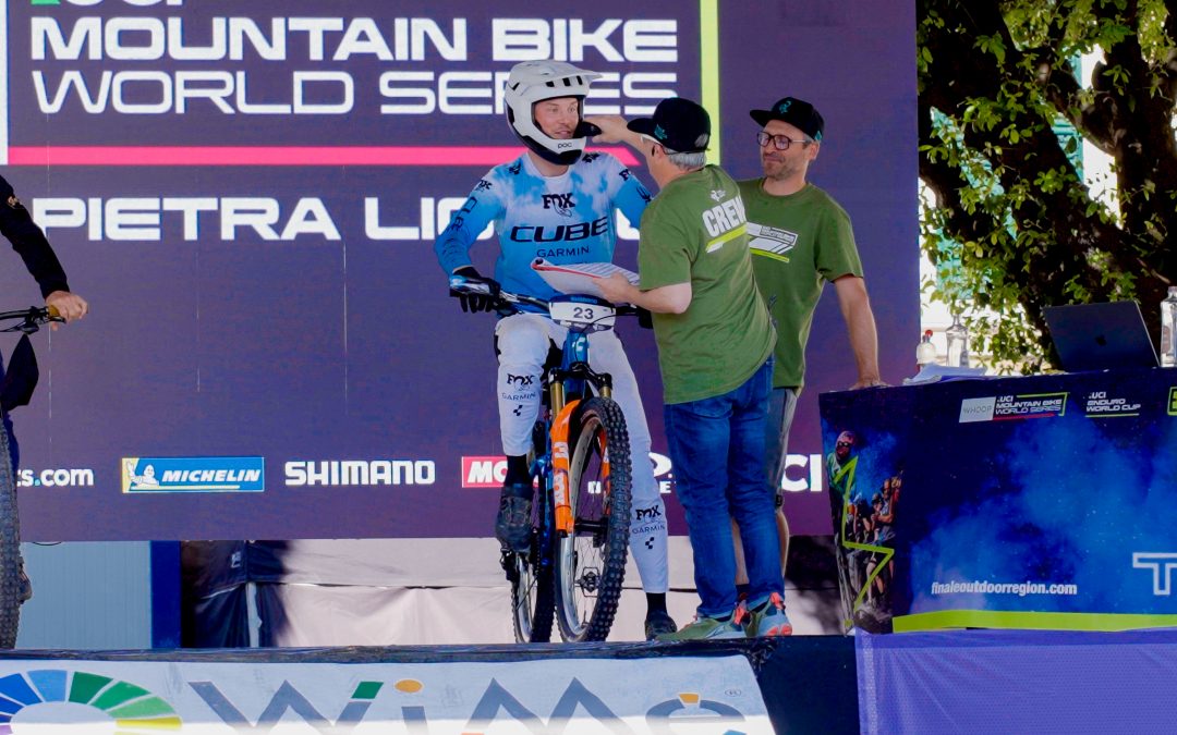 Ein Mountainbiker in blau-weißem Outfit und Helm wartet an der Startlinie und unterhält sich mit zwei Mitarbeitern der Veranstaltung. Auf dem Hintergrund steht "Mountain Bike World Series Pietra Ligure".