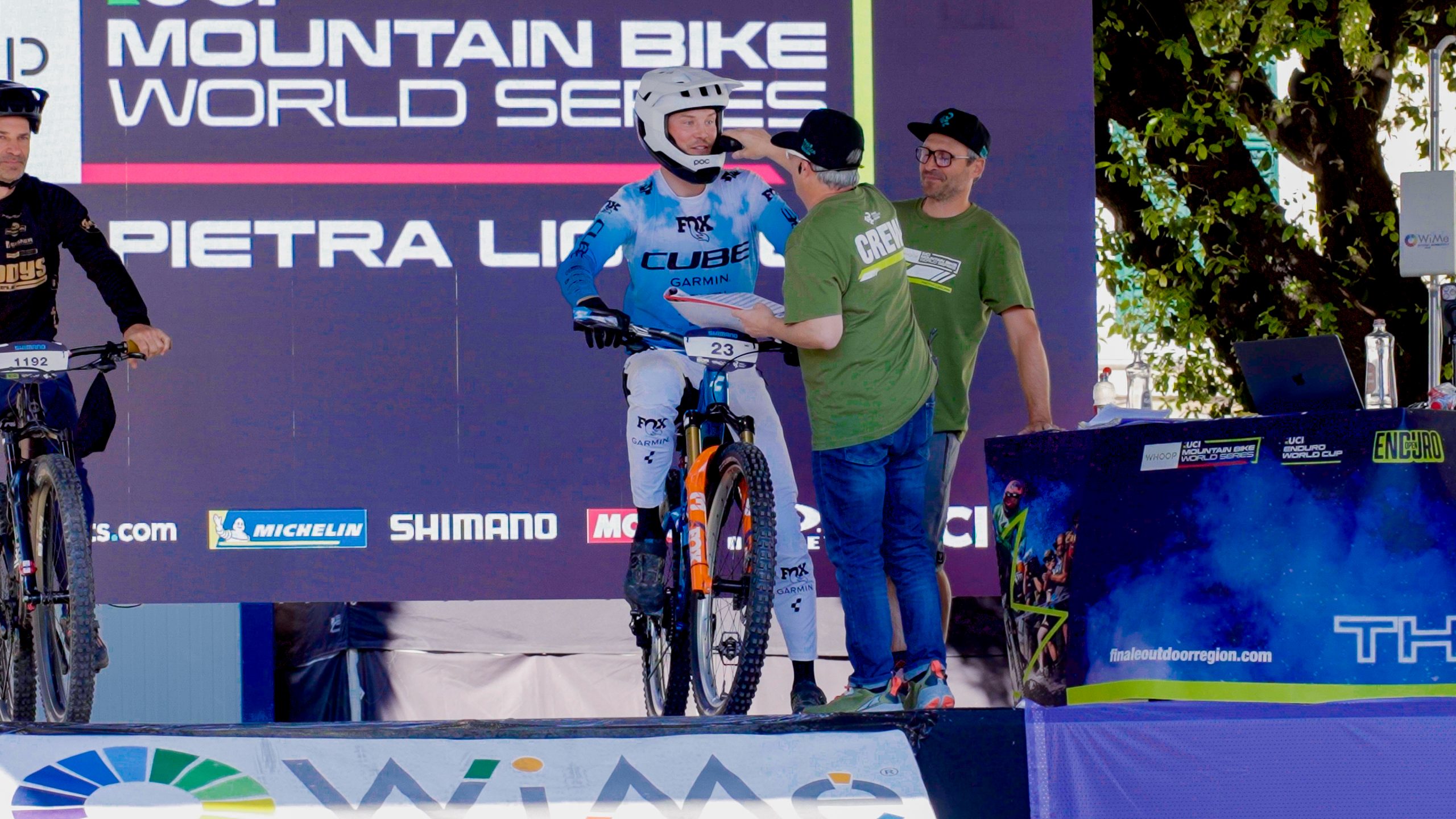 Ein Mountainbiker in blau-weißem Outfit und Helm wartet an der Startlinie und unterhält sich mit zwei Mitarbeitern der Veranstaltung. Auf dem Hintergrund steht "Mountain Bike World Series Pietra Ligure".