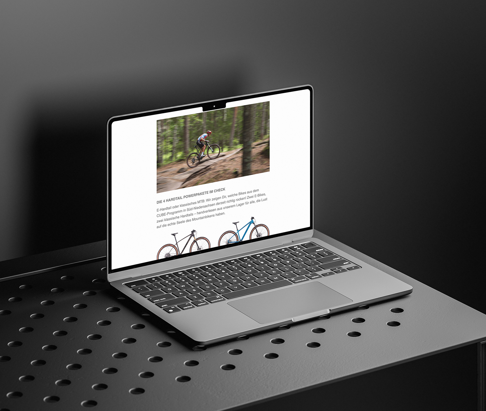Ein Laptop auf einer perforierten Metalloberfläche zeigt eine Webseite mit Bildern und Beschreibungen von Fahrrädern an, mit einem unscharfen Waldhintergrund in einem Bild. Die Umgebung hat eine schlichte, moderne, monochrome Ästhetik.