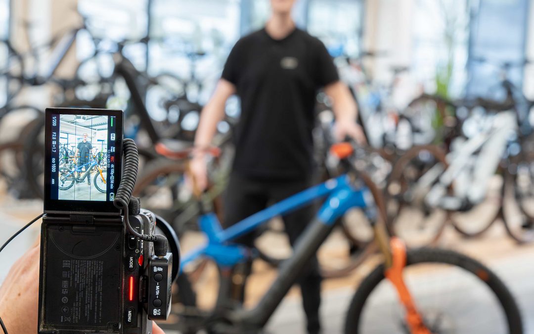 Eine Person hält eine Kamera in der Hand und fokussiert einen Mann, der neben einem blau-orangenen Fahrrad in einem Fahrradgeschäft steht. Auf dem Display der Kamera sind der Mann und das Fahrrad scharf abgebildet, während der Hintergrund unscharf ist.