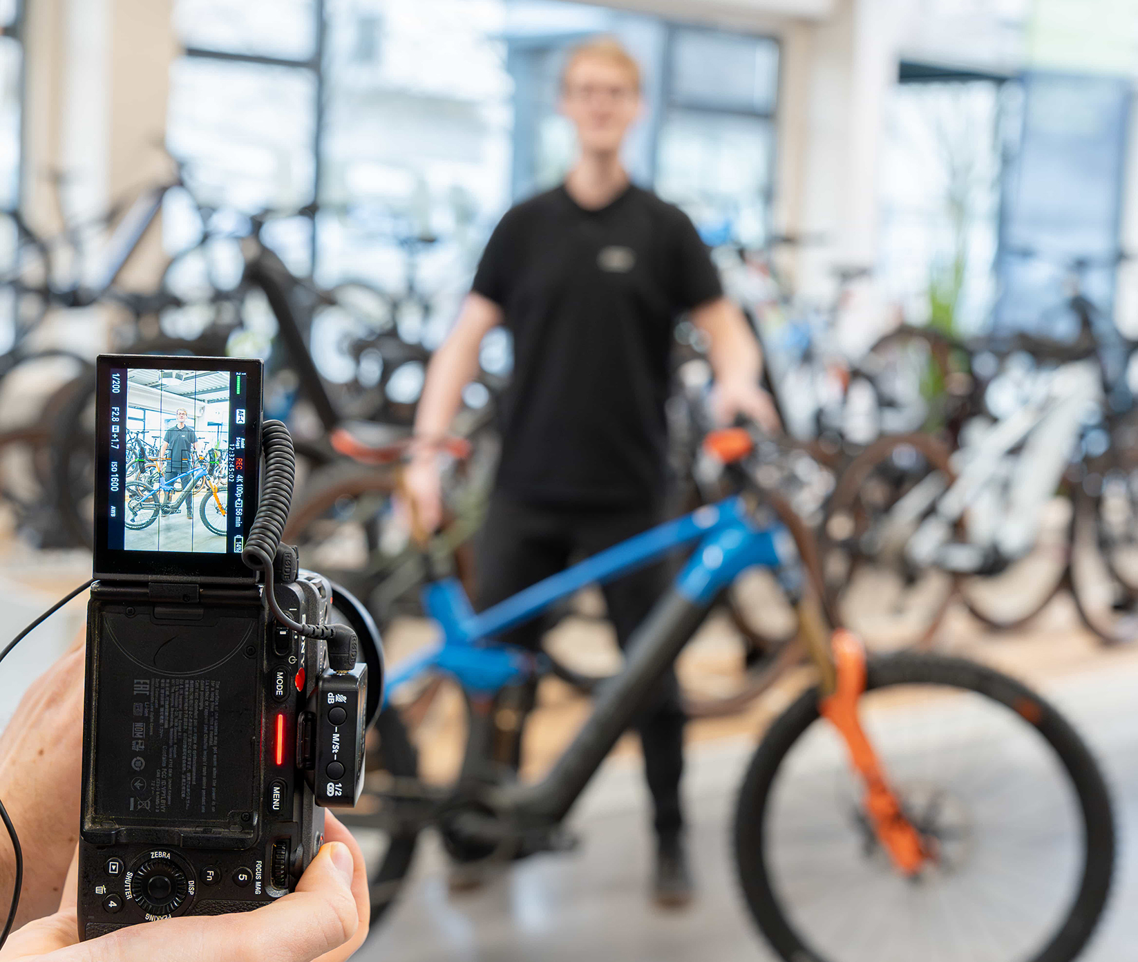 Eine Person hält eine Kamera in der Hand und fokussiert einen Mann, der neben einem blau-orangenen Fahrrad in einem Fahrradgeschäft steht. Auf dem Display der Kamera sind der Mann und das Fahrrad scharf abgebildet, während der Hintergrund unscharf ist.