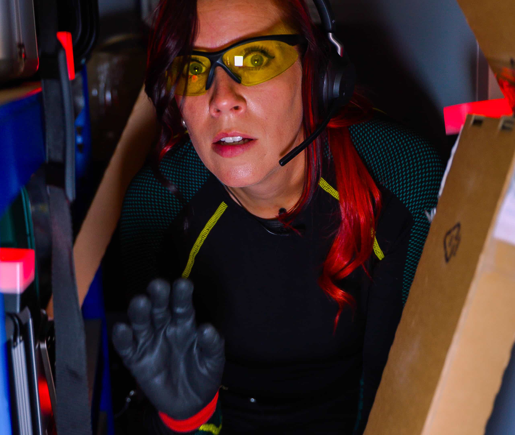 Eine rothaarige Frau mit gelber Schutzbrille, schwarzem Headset und schwarzen Handschuhen hockt zwischen Kisten und hebt die behandschuhte Hand, als würde sie ein Zeichen geben oder sich bereit machen, sich zu bewegen.